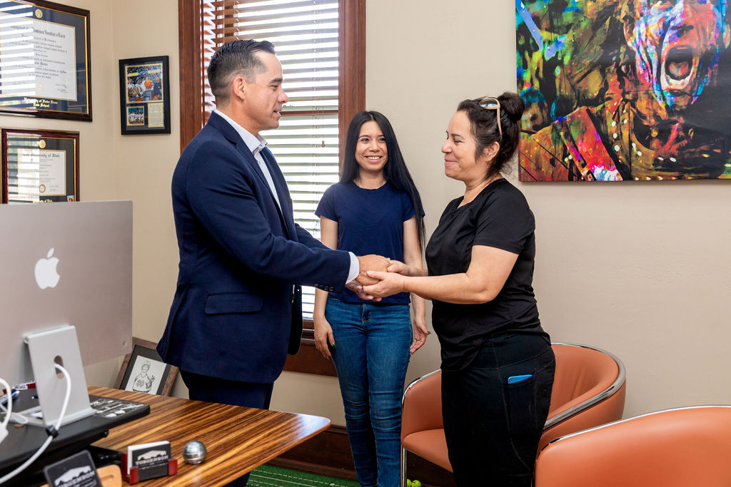 A man in a suit shakes hands with a woman, while another woman stands smiling between them in an office.