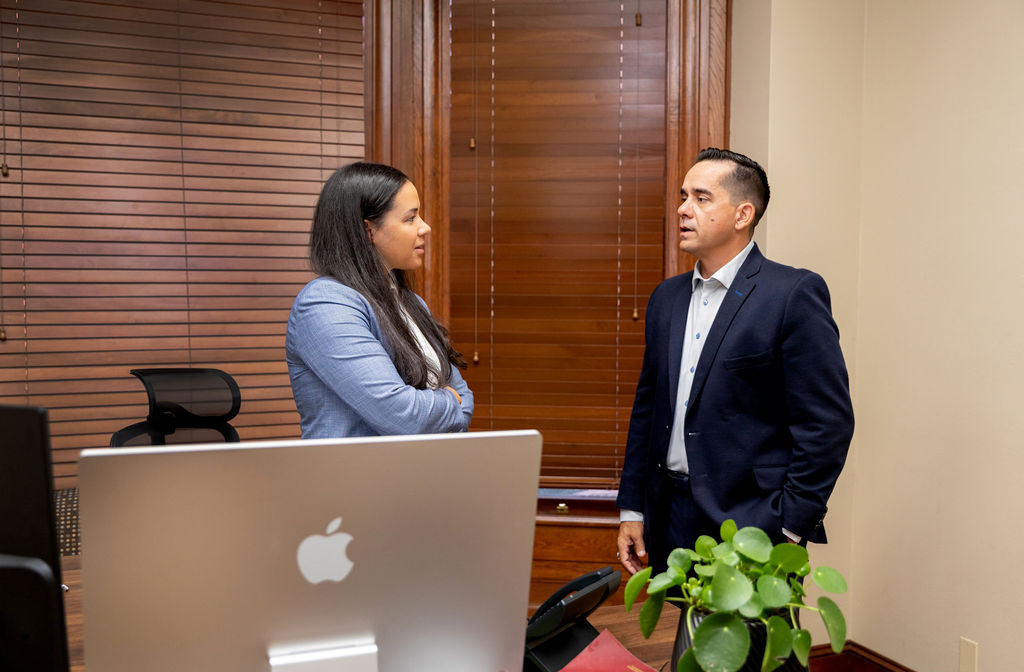 A woman in a blue blazer and a man in a navy suit standing in an office talking to each other for our About Us page.