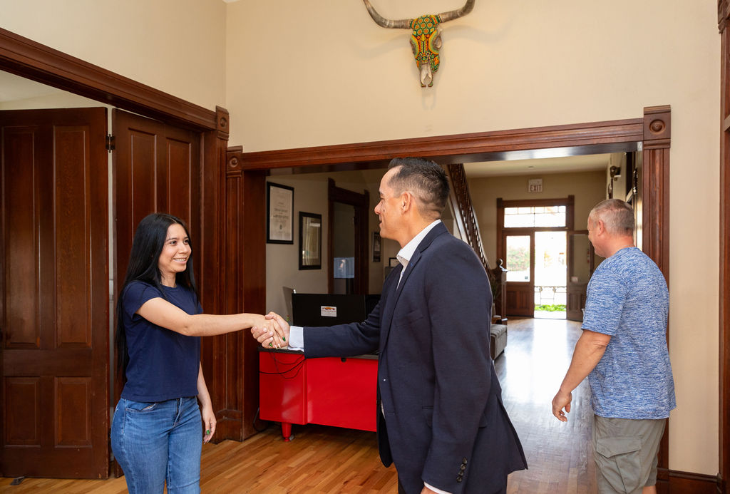 A man in a T-Bone suit shakes hands with a woman in a blue t-shirt and jeans in a building lobby, while a man in a blue patterned shirt walks away in the background.