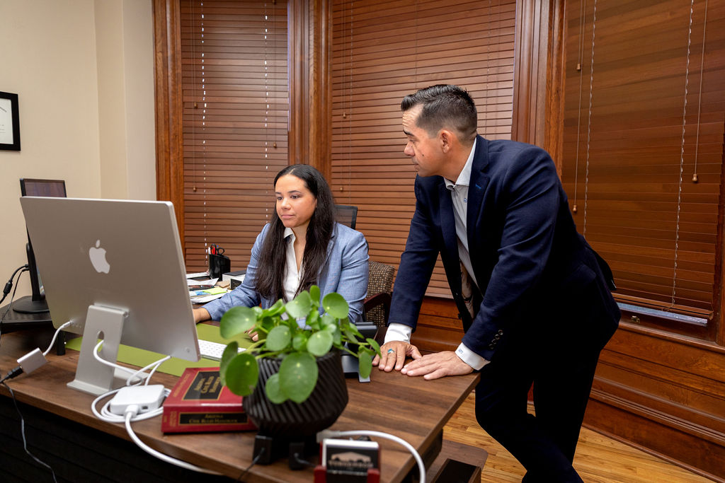 A man in an ATV-branded suit leans over a desk to consult with a woman working on a desktop computer in an office.