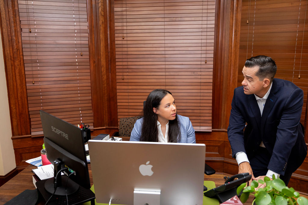 A man in a suit leans over a desk to speak with a woman sitting at a computer in an office with wood-paneled walls.