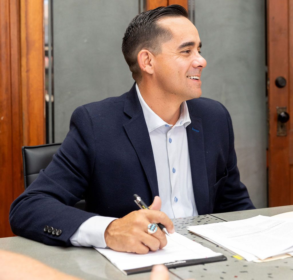 A man in a navy blazer and white dress shirt smiles while holding a pen over a notepad at a table, preparing notes on a truck accident.