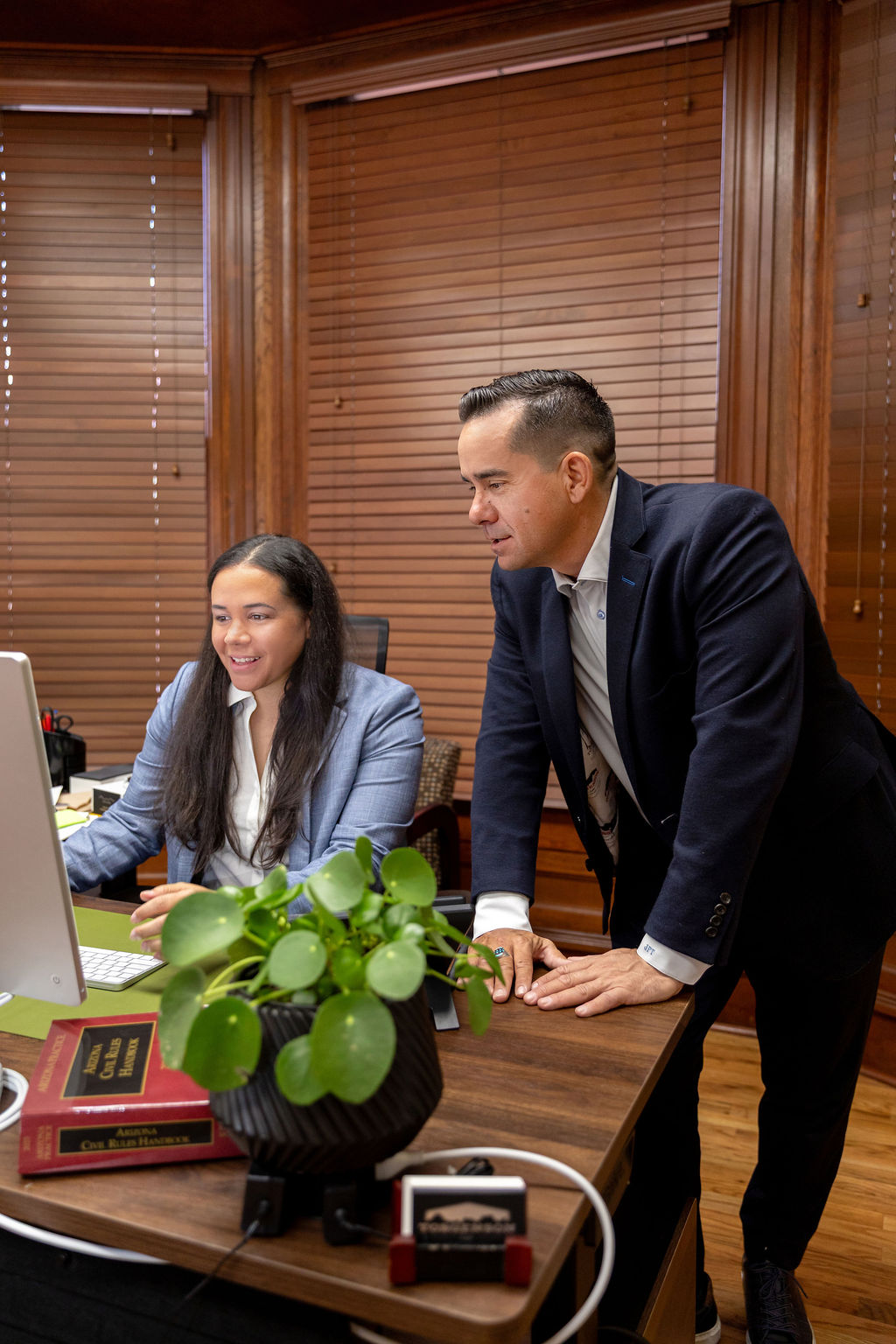 A man in a suit leans over a desk to consult with a woman in a blue blazer, who is looking at a computer screen showing information about a car accident.