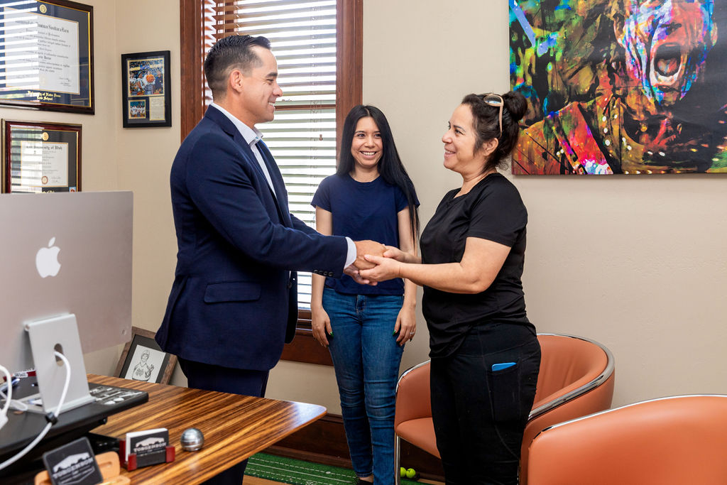 A man in a suit shakes hands with a woman in an office as another woman stands behind them smiling.