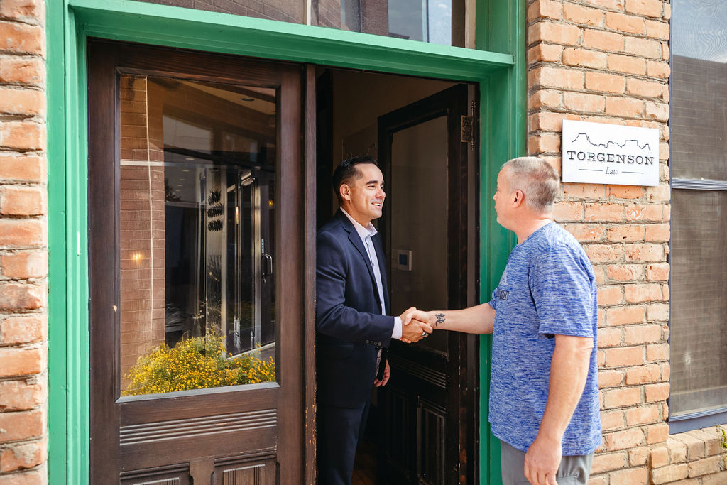 A man in a suit shakes hands with T-Bone in a blue t-shirt in the doorway of a brick building with a sign that reads Torgenson Law.