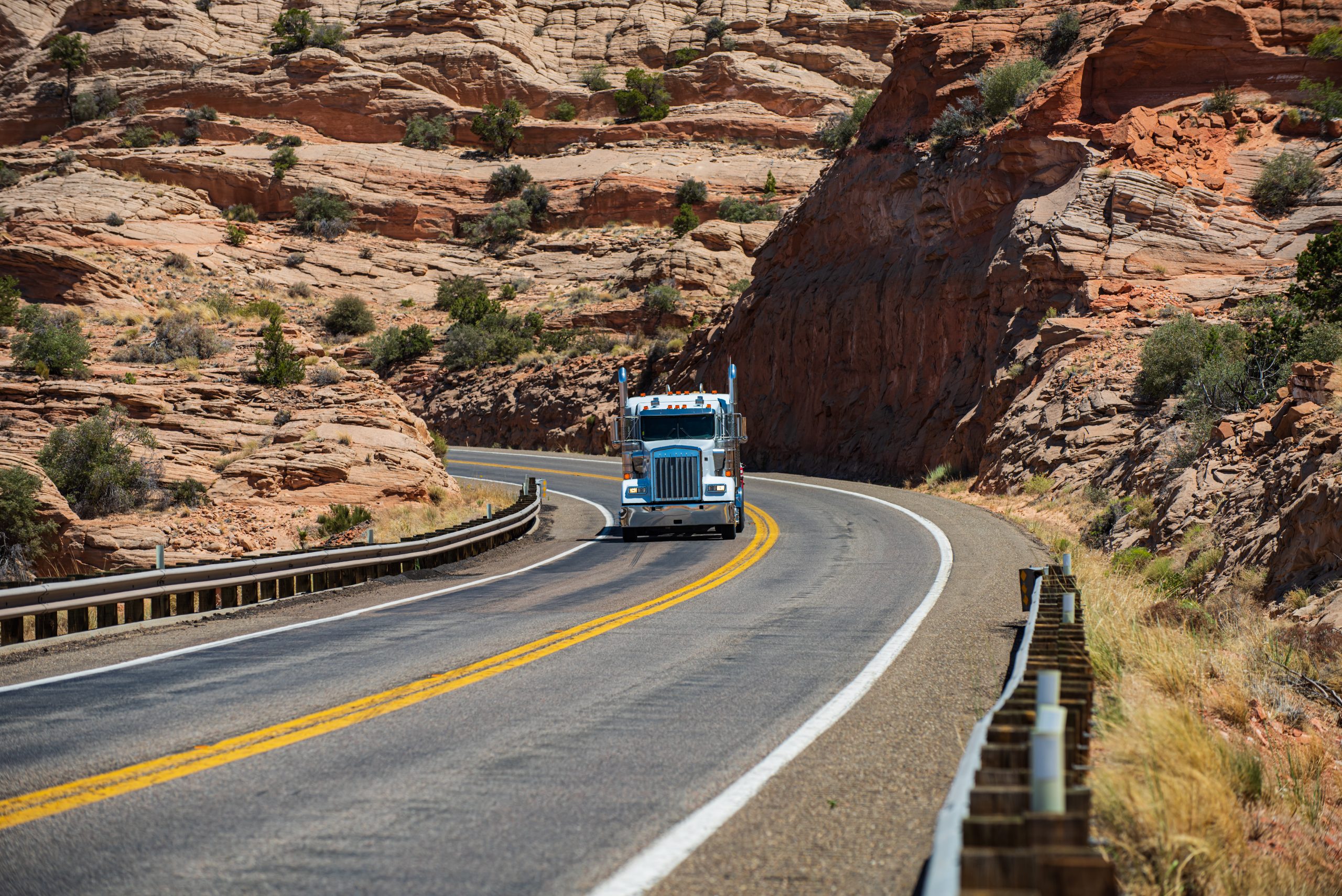 A semi-truck involved in an accident drives along a winding road through a rocky desert landscape.