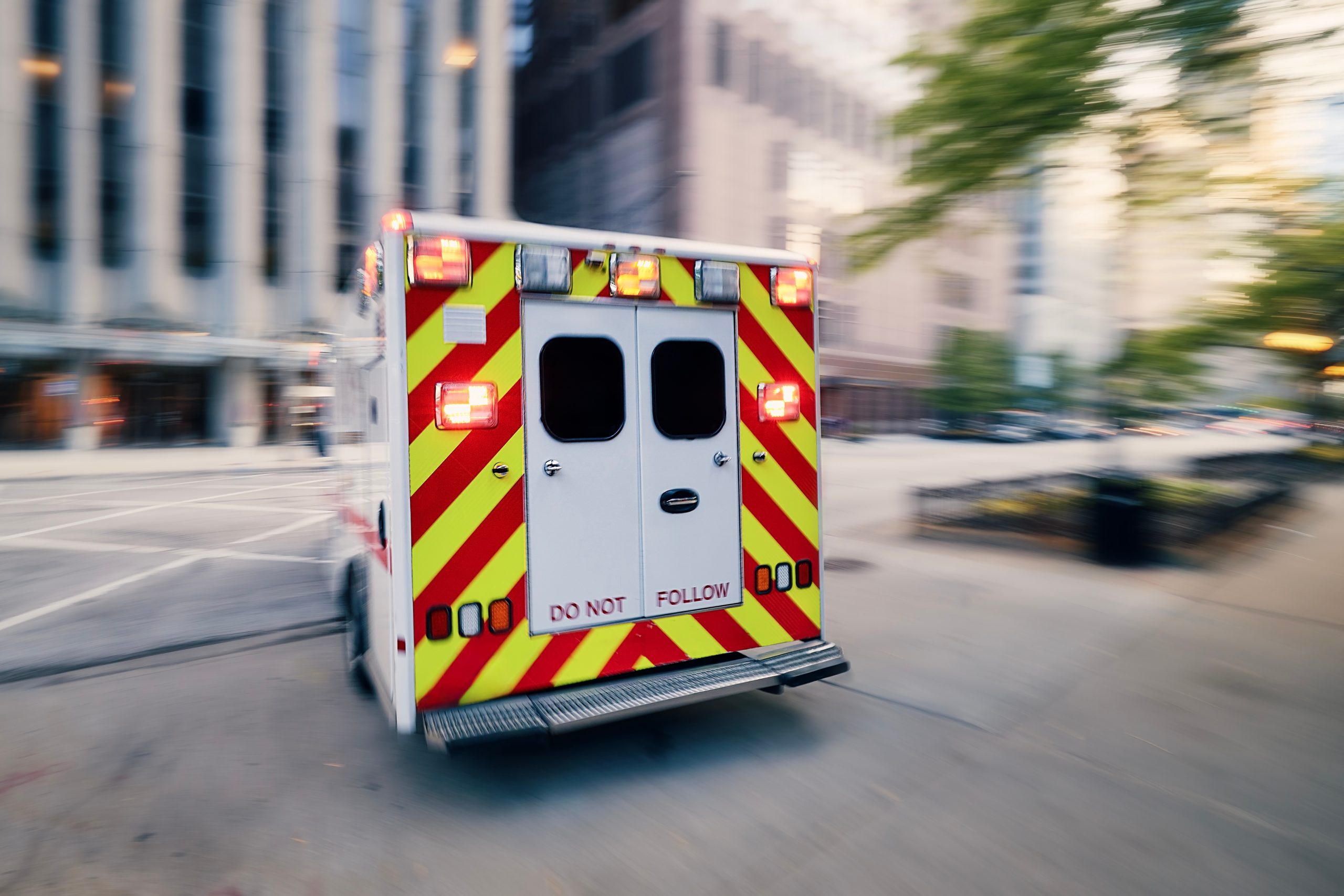 An ambulance with flashing red lights drives quickly down a city street, likely responding to a truck accident.